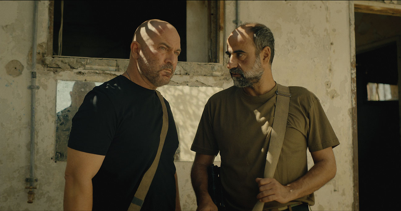 "Two serious men with shoulder bags stand in front of a weathered, sunlit building, partially shaded by window light inside a worn, possibly abandoned environment."